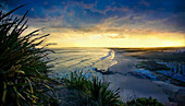 Blick auf den ruhigen Strand, der unter einem goldenen Sonnenuntergangshimmel auf den Ozean trifft, mit üppigem grünen Laub im Vordergrund, Redhead Beach, Newcastle, Australien.