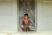 Mirsharai, Bangladesh - 21 October 2019: View of a smiling boy crouched in front of a weathered wooden door, light catching the textured wall behind him.