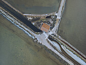 Aerial view of geometric salt pans intersected by narrow channels, creating a mosaic of earthy tones, near a quaint, red-roofed structure, Gruissan, Occitanie, France.