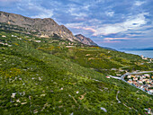 Luftaufnahme der üppig grünen Hügel, die zur Adria hin abfallen, wo Terrakotta-Dächer auf das azurblaue Wasser unter einem pastellfarbenen Himmel treffen, Podgora, Gespanschaft Split-Dalmatien, Kroatien.
