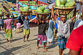 Dhaka, Bangladesch - 08. April 2023: Blick auf Arbeiter, die Wassermelonen in Körben auf ihren Köpfen tragen, eine lebhafte Szene von Handel und Arbeit in der Nähe des Flussufers.
