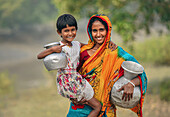 Bogura, Bangladesch - 13. Dezember 2019: Blick auf eine Mutter und ihr Kind, die in leuchtenden Farben gekleidet sind und einen zärtlichen Moment teilen, während sie in einer ländlichen Landschaft Wassertöpfe tragen.