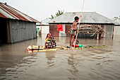 Kurigram, Bangladesch - 21. Juni 2022: Blick auf eine Mutter und ihr Kind, die auf einem behelfsmäßigen Floß durch die Fluten fahren, vorbei an überschwemmten Häusern, deren Wellblechdächer sich im trüben Himmel spiegeln.