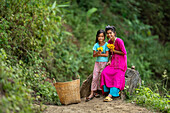 Bogura, Bangladesh - 04 December 2023: View of two young girls, one in vibrant pink, the other in pale blue, seated amidst lush greenery, holding yellow flowers.