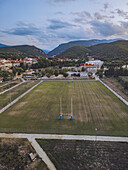 Aerial view of a vibrant green sports field with blue goalposts nestled between terracotta rooftops and verdant hills under a pastel sky, Prades, Occitanie, France.