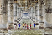 Bogura, Bangladesch - 19. Oktober 2017: Blick auf Kinder, die im Fluss unter den kahlen, imposanten Betonpfeilern einer Brücke waten, deren spielerische Energie einen lebhaften Kontrast zur Brücke bildet.