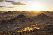 View of golden sun sinking over the stark, dramatic landscape of black volcanic hills and sandy valleys, casting long shadows, Bawiti, Giza Governorate, Egypt.