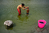 Bogura, Bangladesch - 03. März 2017: Blick auf einen zärtlichen Moment, in dem eine Frau sanft ein Kind im Fluss wäscht, während in der Nähe Wäsche gewaschen wird, was den einfachen Rhythmus des Lebens widerspiegelt.