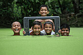 Mirsharai, Bangladesch - 08. Juli 2020: Blick auf Kinder, die freudig aus einem leuchtend grünen Teich auftauchen, eingerahmt von einem alten Fernsehgerät vor einer Kulisse aus üppigem Laub.