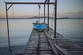 View of a weathered blue boat sits poised at the end of a wooden pier, reflecting the soft pastel hues of the sunset sky, Etang de Thau, Occitanie, France.