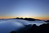 Blick auf ätherische Wolken, die sich über die dunklen, zerklüfteten Gipfel wälzen, während die Sonne in einem Feuer aus Orange und Gelb aufgeht und eine Szene von erhabener Schönheit schafft, Funchal, Madeira, Portugal.