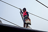 Chittagong, Bangladesh - 28 November 2019: View of a young boy standing atop a weathered boat, his small hand gripping a rope against the pale sky.