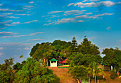 Sajek, Bangladesh - 27 January 2019: View of a lush green hill crowned with vibrant trees, a small white building, and the fluttering Bangladeshi flag against a bright blue sky.