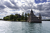 View of Boldt Castle's imposing stone structure rises from the water, framed by lush greenery and a sky full of dramatic clouds, St Lawrence River, New York, United States of America.