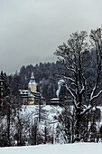 View of snow-covered ground and trees with a beige building and a tower nestled among the hills on a cloudy day in Almau, Bavaria, Germany.
