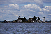 View of a scenic island, hugged by the tranquil waters, hosting a historic lighthouse and quaint buildings under a sky brushed with clouds, St Lawrence River, New York, United States of America.