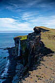 View of a waterfall cascading from rugged cliffs into the dark ocean waters under a vast blue sky, highlighting the raw beauty, Ketubjörg, Iceland.