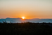 Aerial view of the sun cresting over distant, hazy mountains, casting a warm golden glow across the landscape, Soppeng, Sulawesi Selatan, Indonesia.
