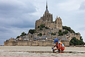 Mont Saint-Michel, France - 19 August 2025: View of a young boy building a sandcastle with the majestic Mont Saint-Michel abbey rising in the background against a cloudy sky.