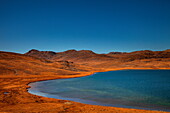 Blick auf das ruhige blaue Wasser im Kontrast zur trockenen, orange-braunen Landschaft unter klarem Himmel, Deosai National Park, Gilgit Baltistan, Pakistan.