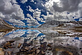 Blick auf das Wasser eines unberührten Sees, in dem sich der strahlende Himmel und die schneebedeckten Berge spiegeln, Steine im Vordergrund, Deosai-Nationalpark, Gilgit Baltistan, Pakistan.