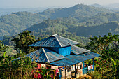 Sajek, Bangladesch - 08. Januar 2018: Blick auf ein leuchtend blaues Holzhaus inmitten üppig grüner Hügel, ein starker Kontrast zu den gedämpften Tönen der entfernten Berge.