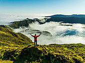 Aerial view of a solitary figure atop a vibrant green peak overlooking a sea of clouds near Lagoa do Fogo, Azores, Portugal.