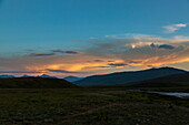 Die Dämmerung färbt den Himmel in Orange- und Blautönen über den sanft geschwungenen grünen Hügeln und den fernen Bergen im Deosai-Nationalpark, Gilgit Baltistan, Pakistan.