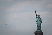 Blick auf die Freiheitsstatue, die sich majestätisch gegen einen wolkenverhangenen Himmel abhebt, während in der Ferne ein Flugzeug vorbeifliegt, ein Symbol für Freiheit und Geschichte, New York, New York, Vereinigte Staaten.