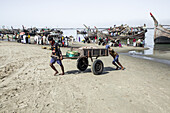 Cox's Bazar, Bangladesh - 29 November 2022: View of men struggling to pull a heavy cart through the sandy beach, with fishing boats clustered along the shoreline under a hazy sky.