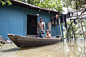 Noakhali, Bangladesch - 07. September 2024: Blick auf einen Mann und ein Kind in einem Holzboot vor einem blauen Wellblechhaus, das von den Fluten umgeben ist.