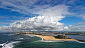 Luftaufnahme der Küstenlinie, wo der Ozean auf den Sandstrand trifft, mit dem Leuchtturm Nobbys Head, der unter einem dramatischen Himmel Wache hält, Newcastle, NSW, Australien.