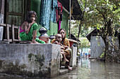Noakhali, Bangladesch - 07. September 2024: Blick auf eine Mutter und ihre Kinder, die auf der Veranda ihres überfluteten Hauses sitzen, während das trübe Wasser die leuchtenden Farben ihrer Kleidung reflektiert.