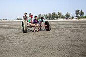 Cox's Bazar, Bangladesch - 08. Februar 2024: Blick auf Kinder, die einen behelfsmäßigen Wagen mit bunten, wiederverwendeten Reifen über den weitläufigen, sonnenverwöhnten Strand ziehen.
