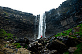 Blick auf eine einsame Gestalt in Rot, die den herabstürzenden Wasserfall inmitten der zerklüfteten, grünen Klippen betrachtet, ein heiterer Moment, Langasandur, Streymoy, Färöer Inseln.