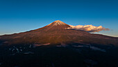 Luftaufnahme des majestätischen Berges Fuji, dessen schneebedeckter Gipfel vom goldenen Licht der Morgendämmerung geküsst wird, eine einsame Wolke, die träge dahinzieht, Fujinomiya, Shizuoka, Japan.