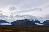View of a glacier tongue snaking its way down from snow-capped mountains under a blanket of moody skies, a symphony of grey and white contrasting with the dark earth, Vik, Iceland.