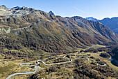 Luftaufnahme einer kurvenreichen Straße, die sich durch die zerklüftete Herbstlandschaft schlängelt, mit Bergen, die sich in der Ferne gegen den klaren Himmel erheben, ein Wandteppich aus Gold und Braun, Poschiavo, Graubünden, Schweiz.