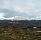 Luftaufnahme von schneebedeckten Gipfeln vor dem Hintergrund eines stimmungsvollen Himmels und einer in Braun- und Grüntönen gehaltenen Landschaft, Loften-Inseln, Nordland, Norwegen.