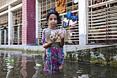 Noakhali, Bangladesh - 02 September 2024: View of a young girl wading through floodwaters, clutching a plastic container, with submerged buildings reflecting in the murky depths.
