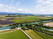 Luftaufnahme der grünen Felder, die unter einem weiten Himmel auf den fließenden Fluss und die Straßen treffen, die eine Mischung aus Natur und Infrastruktur darstellen, Aare, Schweiz.