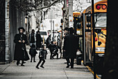 New York, United States - 25 December 2023: View of Orthodox Jewish community members walking near a bright yellow school bus, creating a study in contrasts on a city sidewalk..
