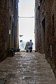 Pienza, Tuscany, Italy - 22 April 2025: View of a couple standing at the end of a narrow stone street, gazing out at the distant blue sea as birds fly overhead.