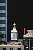 Blick auf einen mit einem goldenen Kreuz gekrönten Kirchturm, der sich zwischen einem eleganten Glas-Wolkenkratzer und einem klassischen Backsteingebäude erhebt, New York, New York, Vereinigte Staaten.