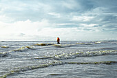 Cox's Bazar, Bangladesh - 11 October 2019: View of a lone figure in an orange jacket stands amidst the turbulent waves of the Bay of Bengal, under a vast, cloud-strewn sky.