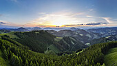 Aerial view of the sun setting over rolling green hills and dense forests, creating a stunning contrast of light and shadow, Teichalm, Steiermark, Austria.