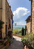 Pienza, Tuscany, Italy - 22 April 2025: View of a narrow stone alleyway lined with potted plants, leading towards a bright, distant valley under a partly cloudy sky.