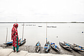 Shyamnagar, Bangladesch - 13. Mai 2023: Blick auf eine Frau in leuchtendem Rot und Grün, die am Ufer steht und den ruhigen, von Fischerbooten gesäumten Fluss überblickt.