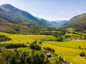 Aerial view of lush green fields meet the rugged mountains, with scattered buildings and a clear blue sky overhead, Stardalen, Vestland, Norway.
