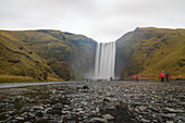 View of majestic Skógafoss waterfall cascading down rugged cliffs, with misty spray and blurred figures adding movement, captured on a cloudy day, Vik, Iceland.